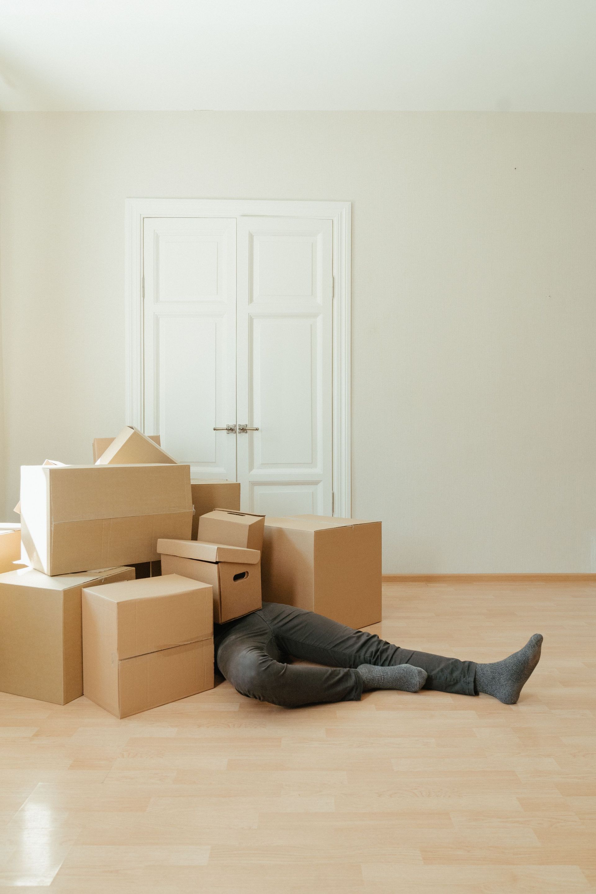 A person lying under a pile of moving boxes in a minimalistic room conveys tiredness from packing.